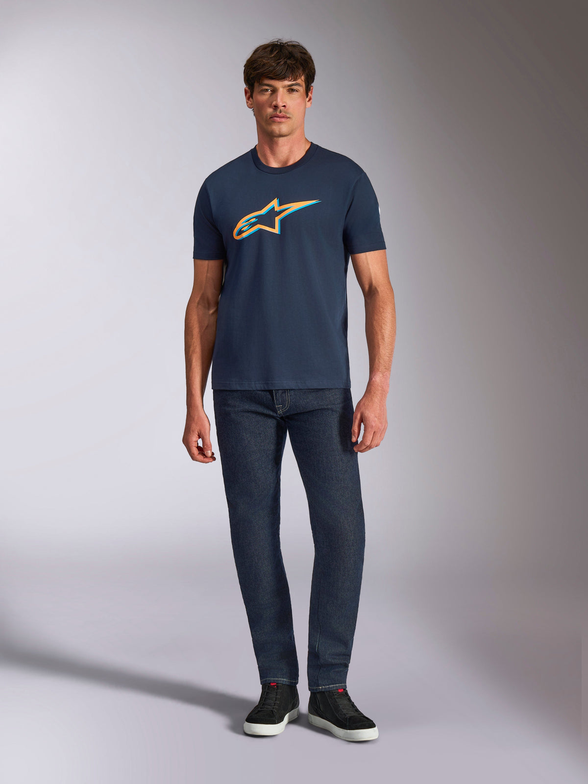 Alpinestars Ageless Shadow CSF Tee, Short Sleeve T-Shirt, Navy Blue, featuring an Orange and Light Blue Ageless Shadow logo, worn by a model with dark denim jeans and black sneakers, casual motorsport-inspired streetwear