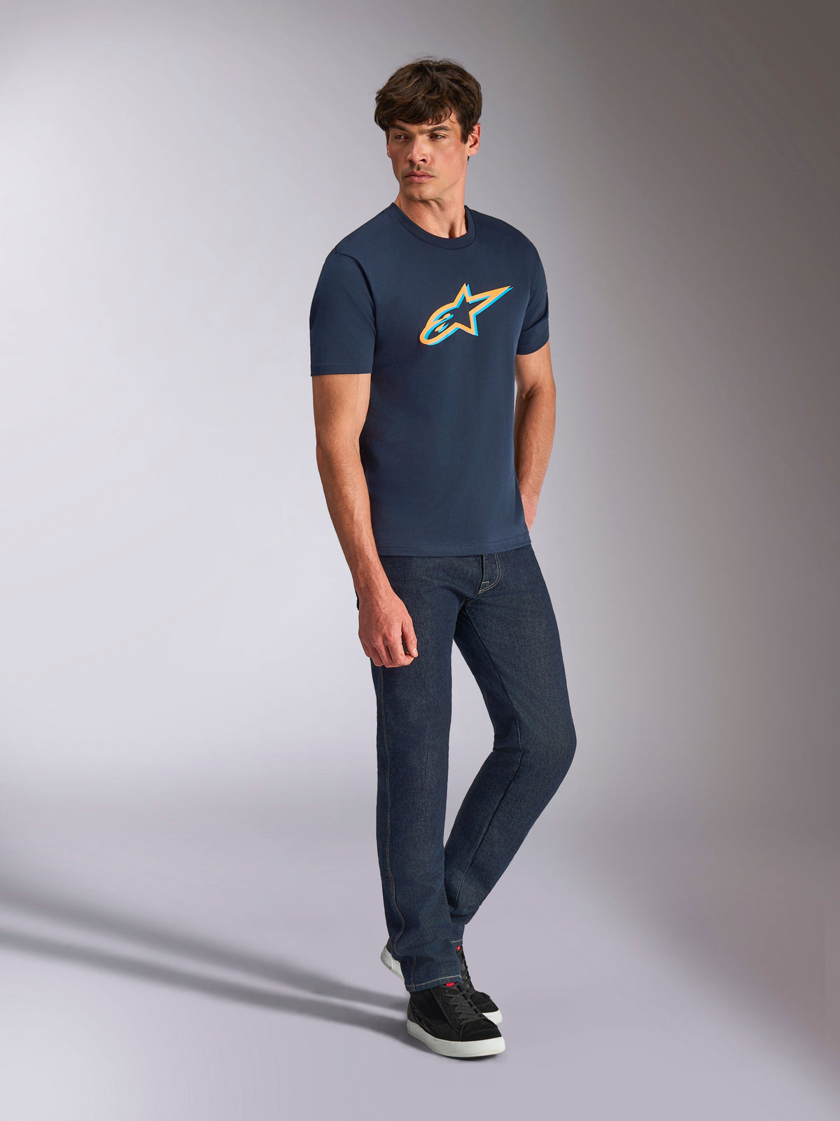 Alpinestars Ageless Shadow CSF Tee, Short Sleeve T-Shirt, Navy Blue with Orange and Teal Shadow Logo, worn by a model with dark denim jeans and black sneakers, casual motorsport streetwear