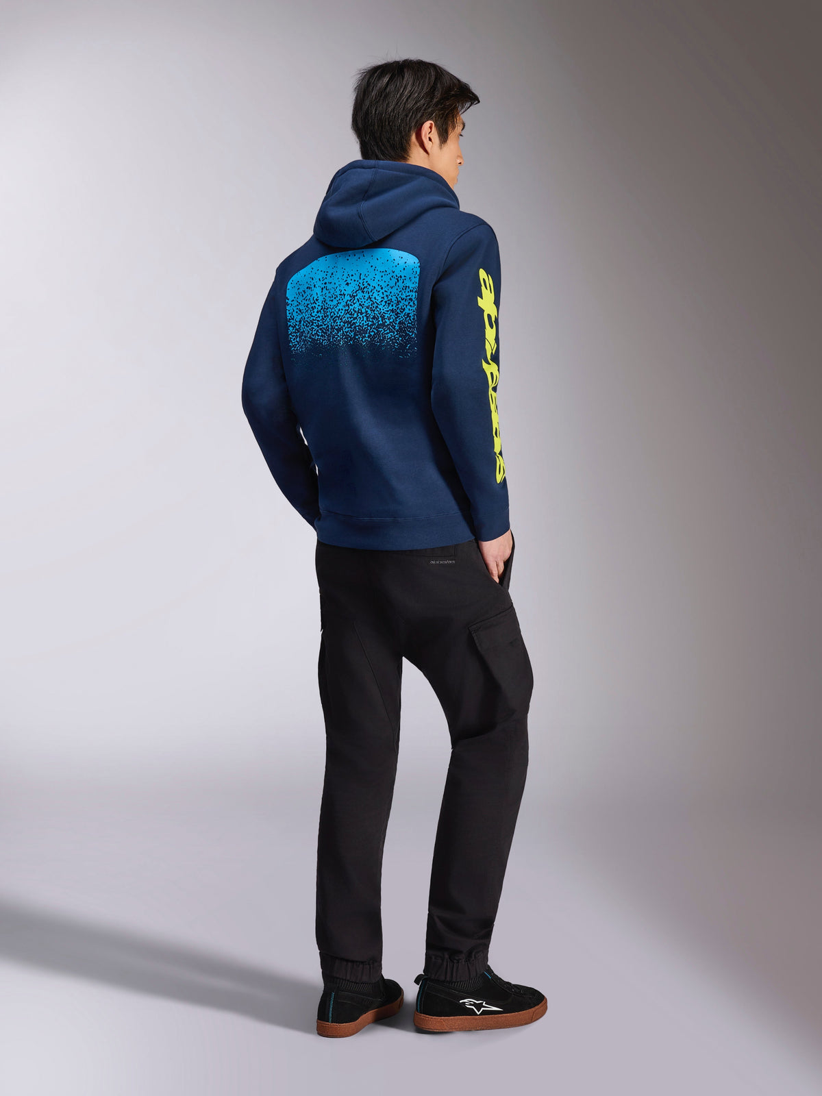 Alpinestars Obsessed Hoodie, Navy Blue casual sweatshirt, back view showing light blue MX gradation graphic and yellow Alpinestars sleeve logo, worn with black pants and sneakers