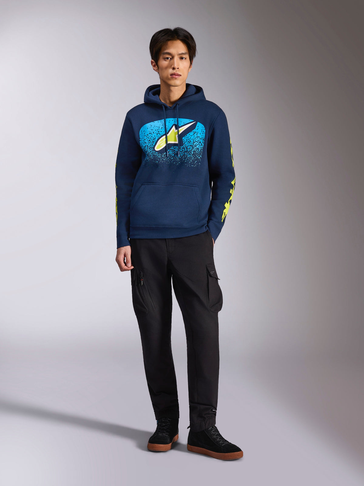 Alpinestars Obsessed Hoodie, Navy Blue pullover hoodie, front Astars logo with blue MX gradation and lime yellow star, lime yellow Alpinestars word sleeve logos, worn by a model with black cargo pants and black sneakers, casual motorsport-inspired streetwear