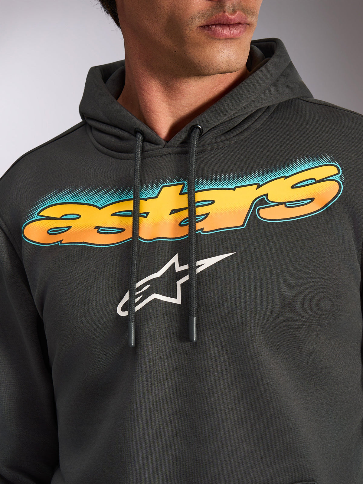 Alpinestars Ellipsoid Hoodie, Gunmetal Gray, close-up of chest featuring curved 'astars' wordmark with yellow-to-orange gradient and teal accents, white Alpinestars star logo, casual motorsport-inspired streetwear fleece