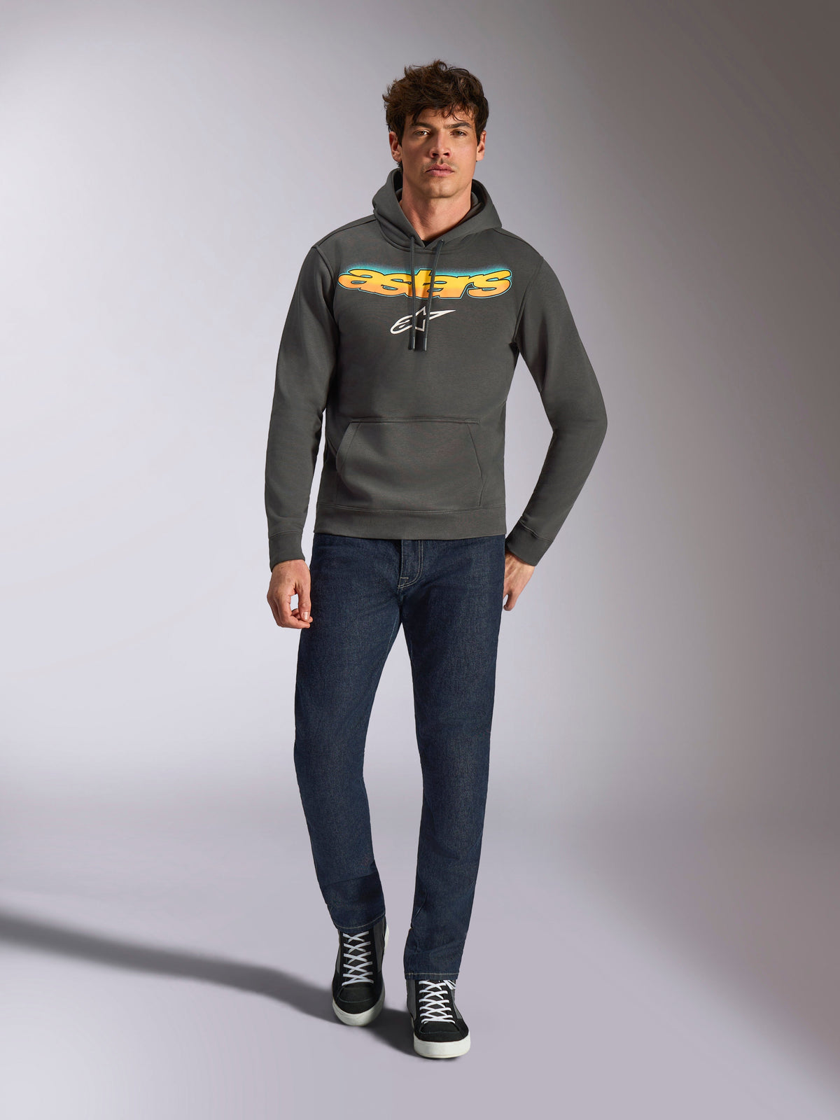 Alpinestars Ellipsoid Hoodie, casual sportswear, gunmetal gray with orange and yellow gradient Astars logo and white symbol, drawstring hood, worn by a male model with dark blue jeans and black sneakers, modern motorsport-inspired streetwear