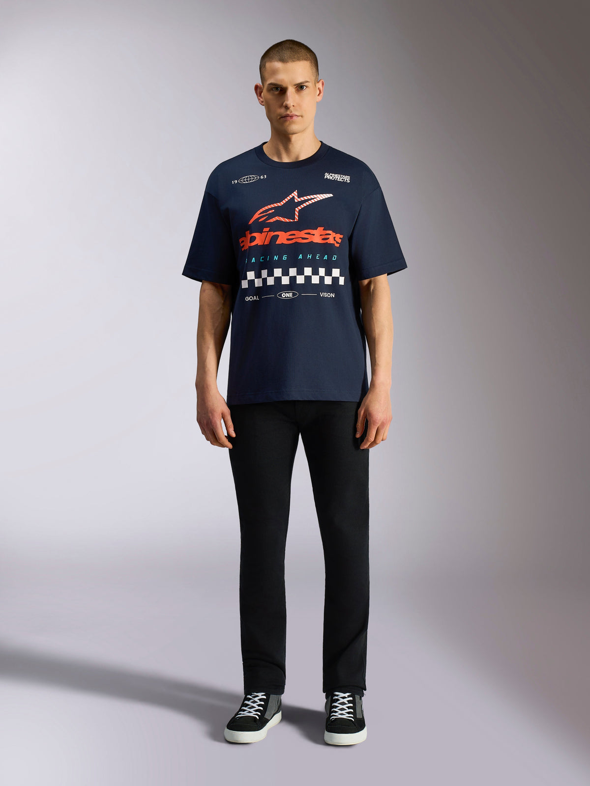 Alpinestars Aplomb Oversized CSF Tee, Short Sleeve Navy Blue T-Shirt, Large Chest Graphic with Orange and Red Gradient Logo and Checkered Pattern, worn by model with black pants and sneakers, Casual Motorsport Streetwear