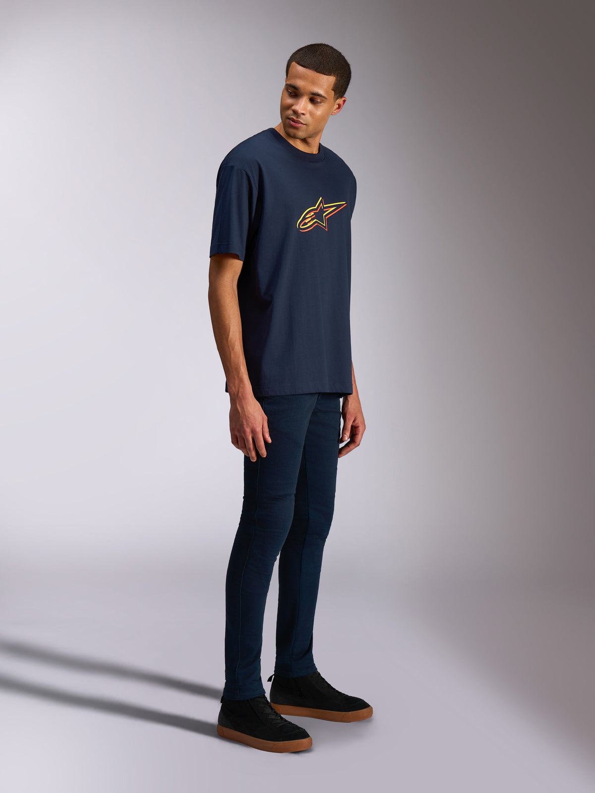 Alpinestars Alter Oversized CSF Tee, Short Sleeve T-Shirt, Navy Blue, Yellow and Orange Astars logo, worn by a model with dark jeans and black sneakers, casual motorsport-inspired streetwear