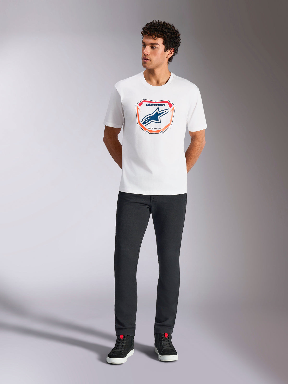 Alpinestars Plate CSF Tee, Short Sleeve T-Shirt, White, featuring a front Astars MX plate graphic with orange and blue accents, worn by a model with black pants and sneakers, Casual Motorsport Streetwear