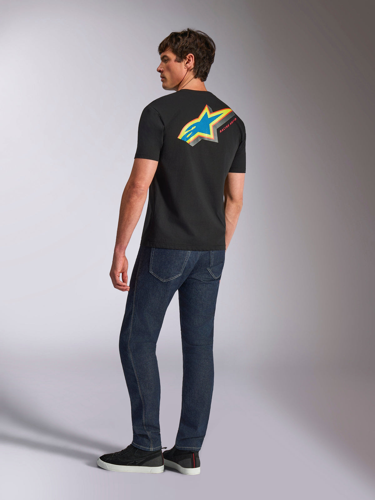 Alpinestars Angler CSF Tee, Short Sleeve T-Shirt, Black, featuring a large vibrant multi-color Astars logo on the back with blue, yellow, and red perforated detail, worn with dark blue jeans, casual motorsport streetwear