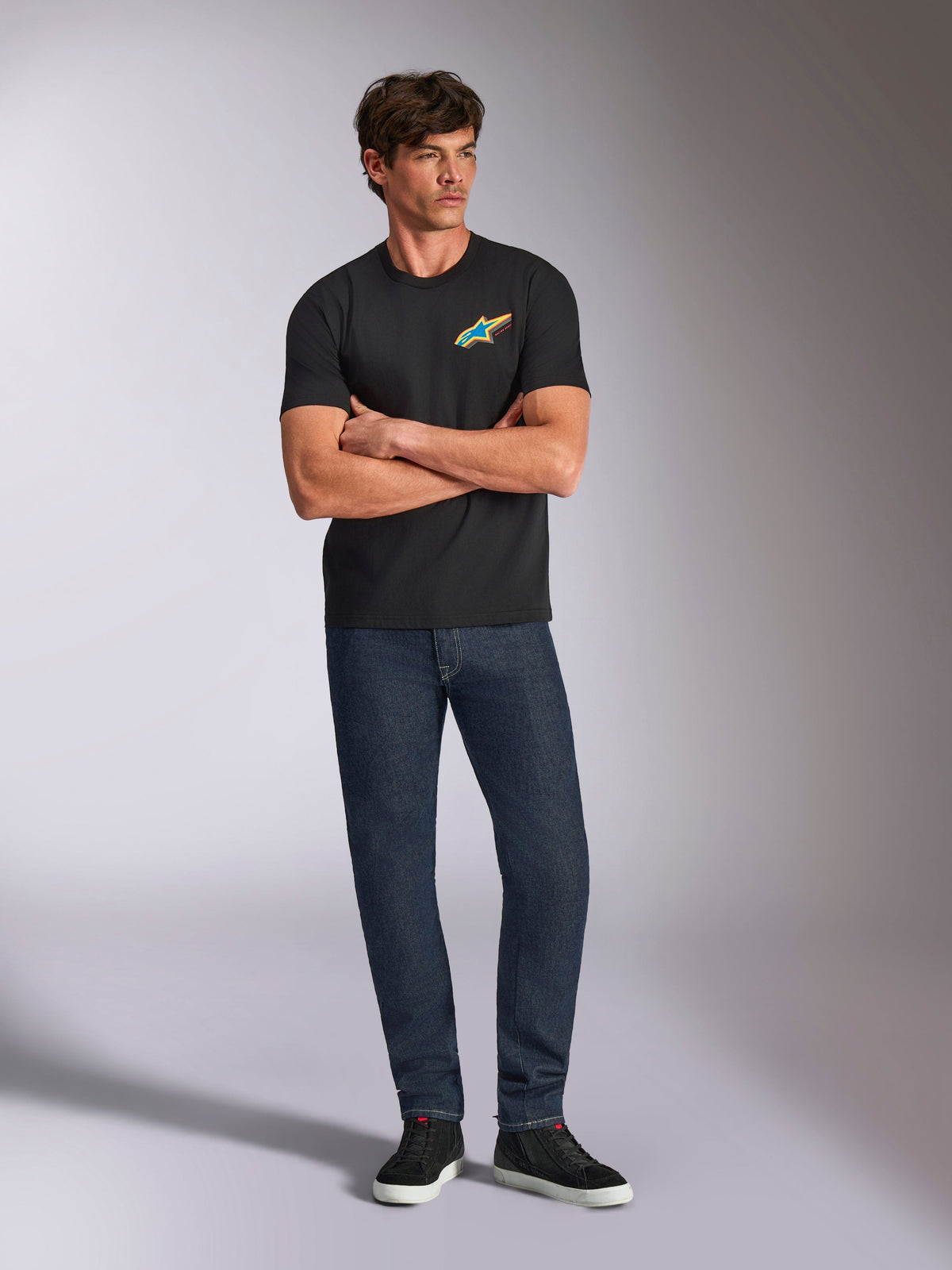 Alpinestars Angler CSF Tee, Short Sleeve T-Shirt, Black, Vibrant Multi-Color Astars Logo on Chest, worn by model with arms crossed, dark denim jeans and black sneakers, casual motorsport streetwear