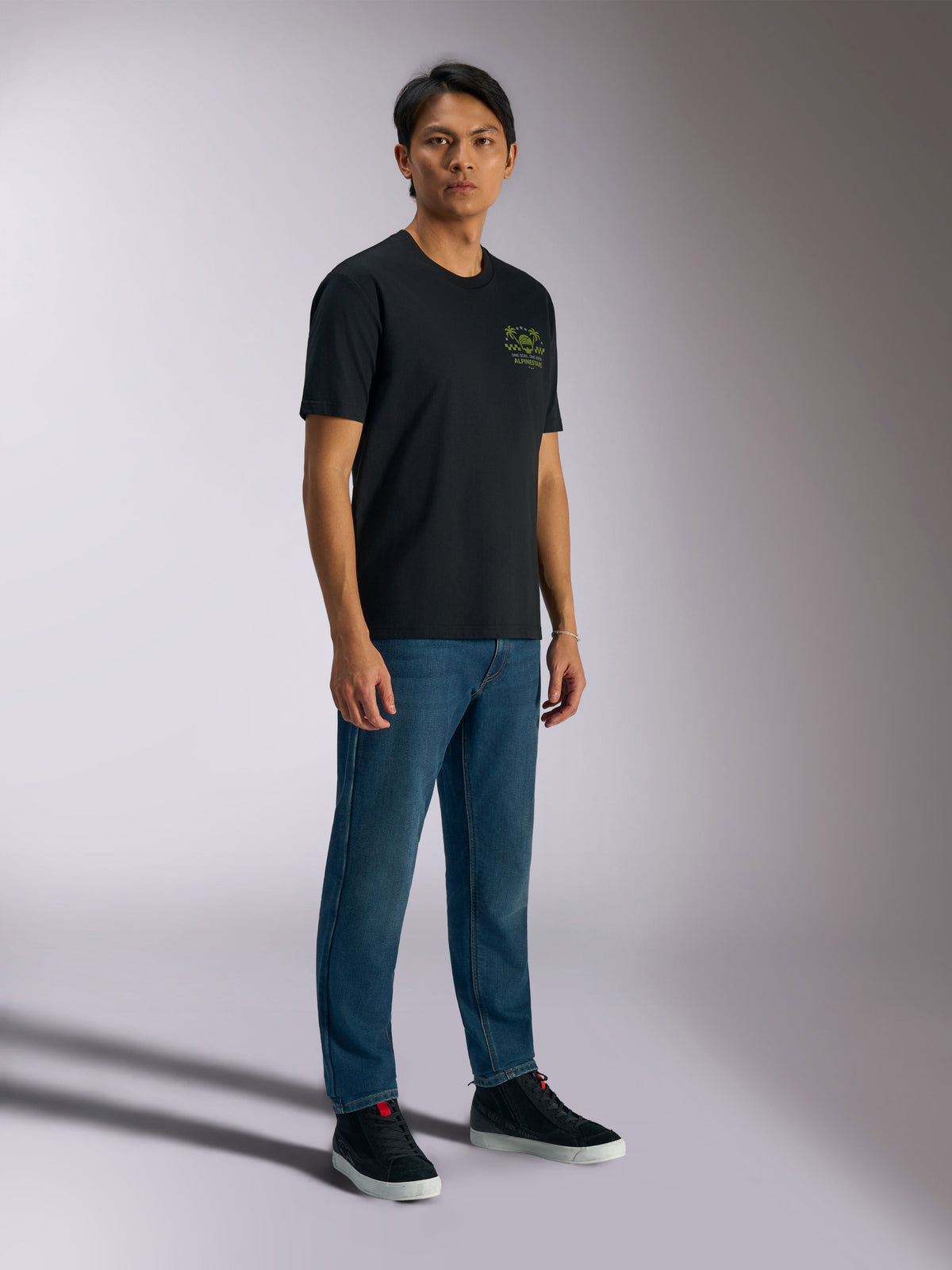 Alpinestars Order CSF Tee, Short Sleeve T-Shirt, Black, Featuring Green Helmet and Palm Tree Logos on Chest, Casual Motorsport Streetwear, Worn with Blue Jeans and Black Sneakers