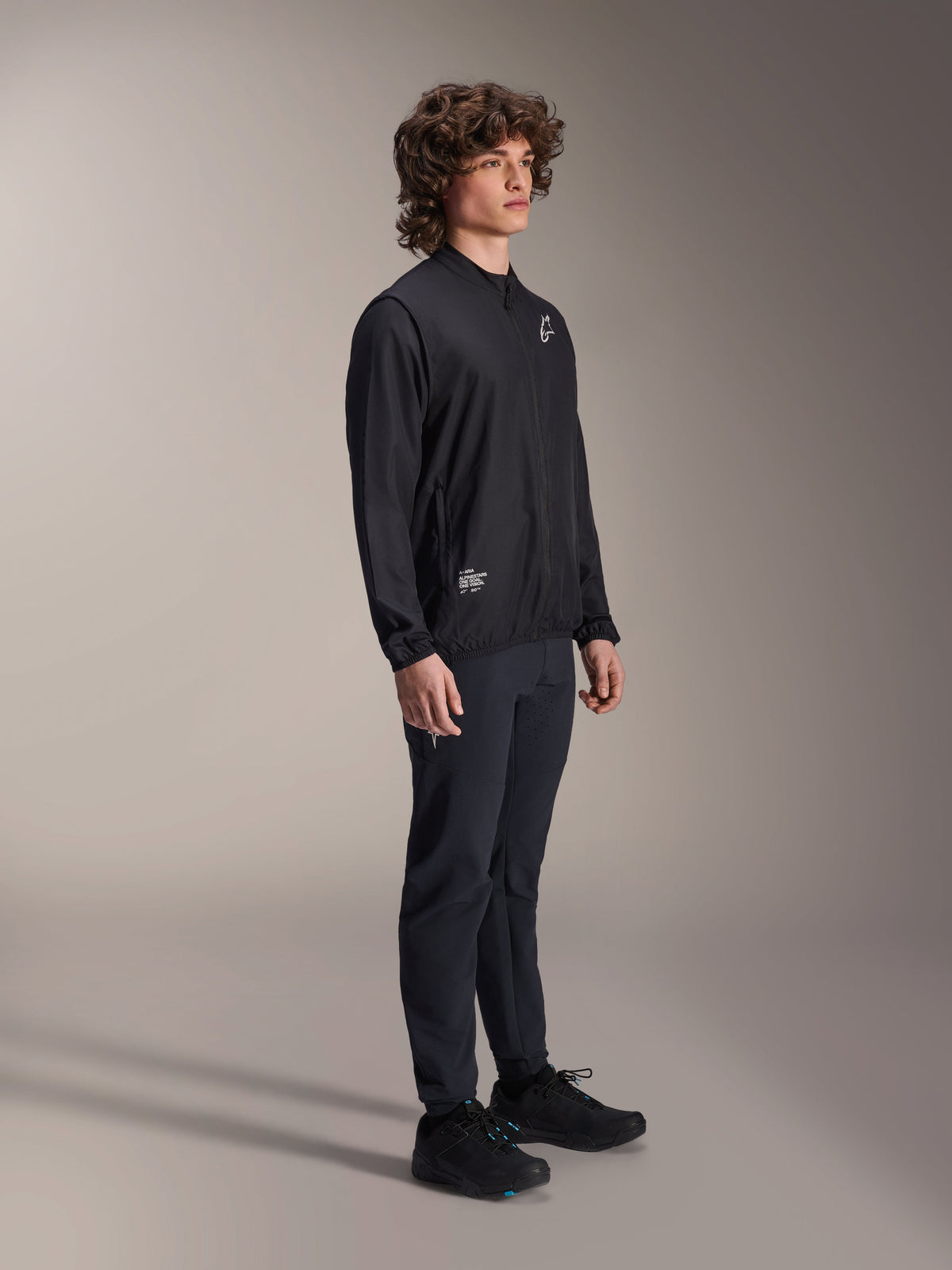 Alpinestars A-Aria Windbreaker Jacket, Black, lightweight wind-resistant cycling jacket worn by a model with curly hair, featuring a white Astars logo on the chest and branding on the lower right, paired with black pants and shoes, performance fit for mountain biking and gravel riding
