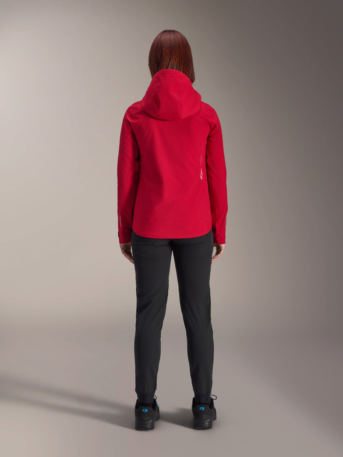 Back view of a woman wearing the Alpinestars Stella A-Dura Elite Waterproof Jacket in Red Fluo, styled with black pants and shoes, featuring a protective hood and dropped tail hem for mountain biking.