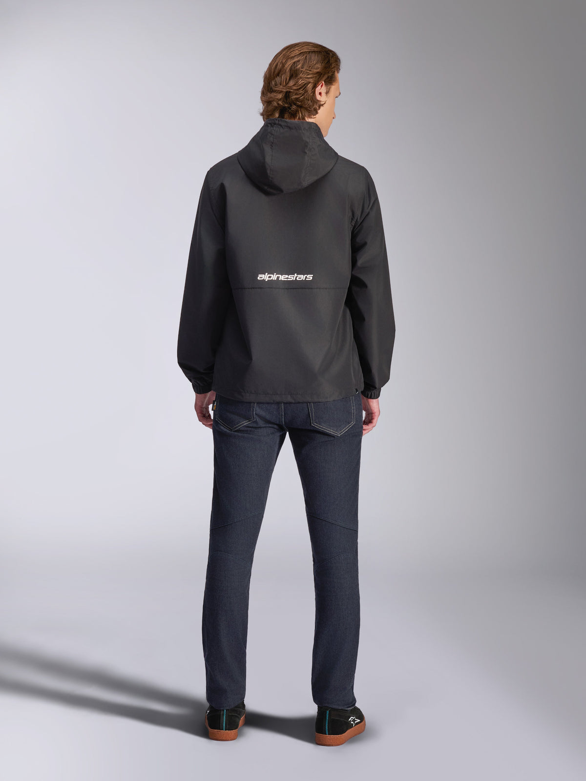 Alpinestars Speeder Chromium Windbreak Jacket, Black, back view with white Alpinestars branding across the middle, hooded style, worn by model with dark denim pants, casual motorsport-inspired jacket