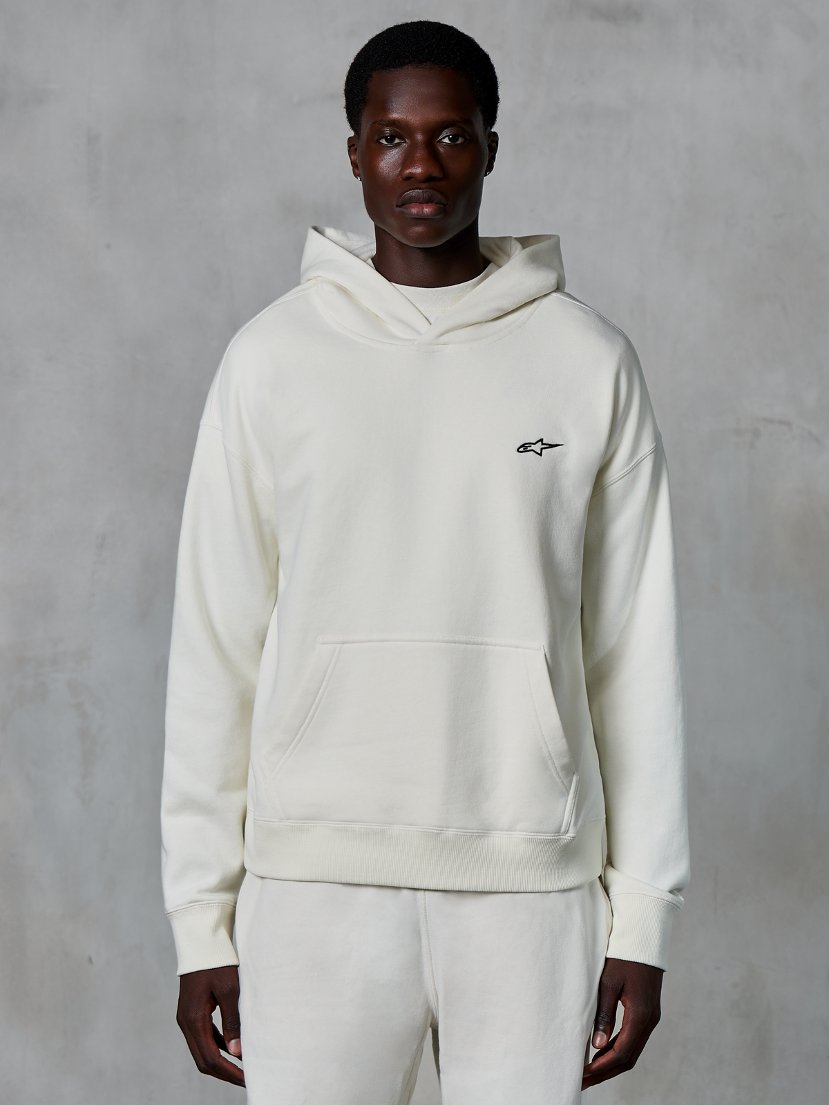 Alpinestars Essentials Ageless Hoodie, off-white, worn by male model, small black logo on chest, front pouch pocket, long sleeves, casual motorsport-inspired streetwear