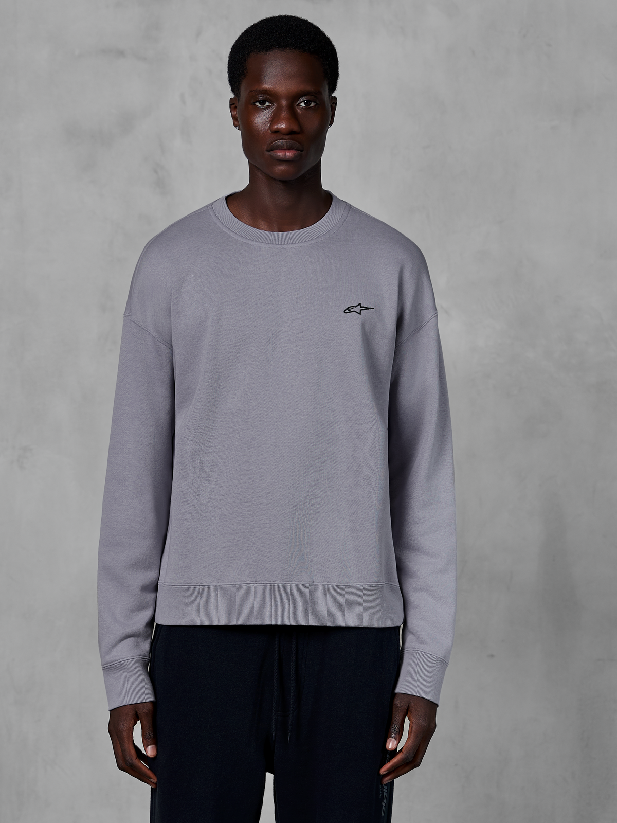 Alpinestars Essentials Ageless Crew, Silver crewneck sweatshirt worn by a model, featuring a small black star logo on the chest, relaxed fit with ribbed cuffs and hem, casual motorsport streetwear