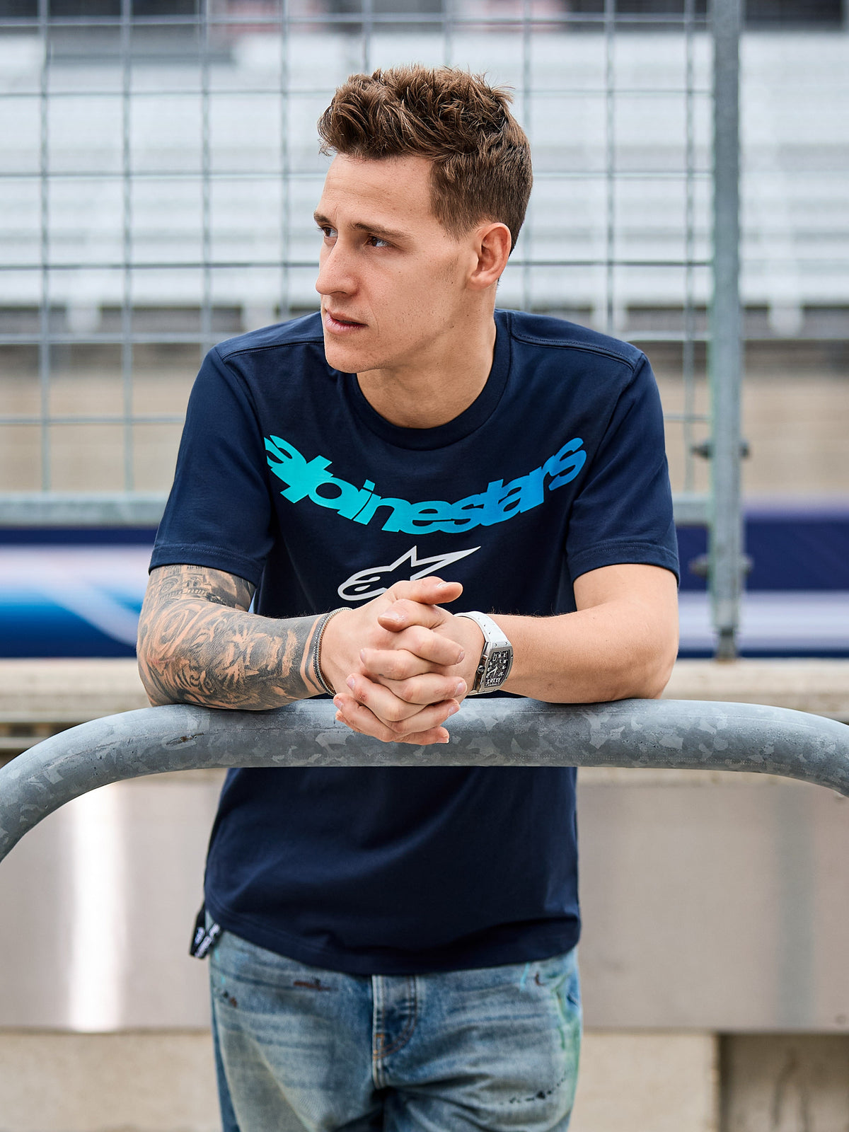 Alpinestars Collared CSF Tee, Short Sleeve T-Shirt, Navy Blue, Teal Alpinestars text and white star logo, worn by a man leaning on a metal railing at a racetrack, casual motorsport streetwear
