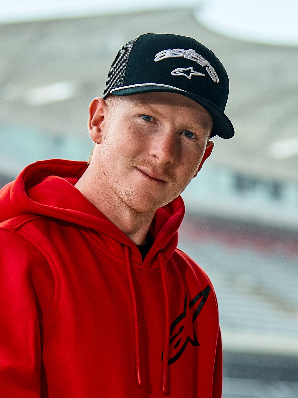 Man wearing a red Alpinestars hoodie and the Agree Trucker Hat in Black, featuring white embroidered Astars logos, a white braided rope detail on the visor, and a breathable mesh back, casual lifestyle setting