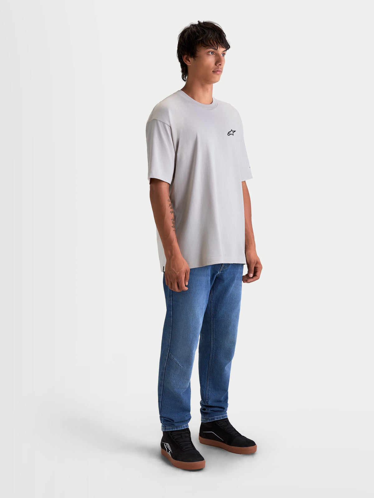 Alpinestars Mandate Oversized CSF Tee, Unisex Short Sleeve Sportswear T-Shirt, Cement Gray, featuring a relaxed oversized fit and small black chest logo. Premium cotton-poly blend construction for casual everyday comfort. Shown on a male model wearing blue jeans and black sneakers.