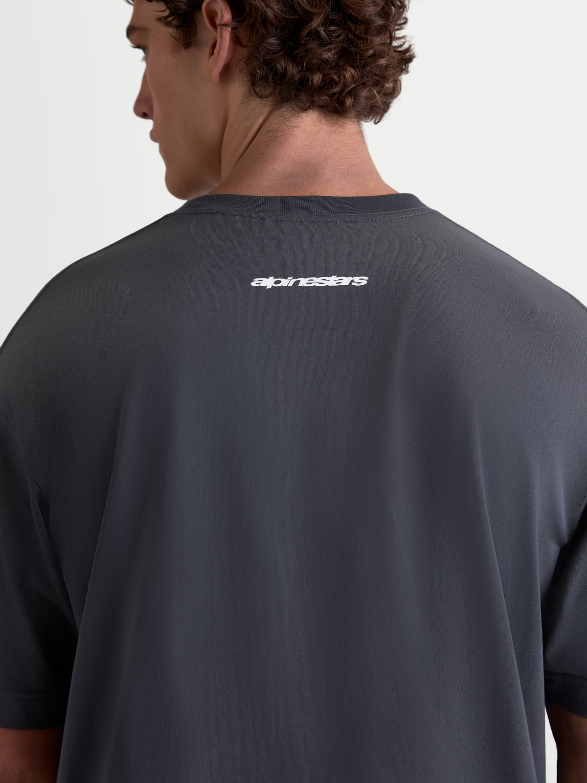 Alpinestars Prelude Oversized CSF Tee, charcoal gray short-sleeve sportswear t-shirt, back view on model featuring a white Alpinestars word logo, relaxed oversized fit for off-bike style, cotton-poly blend fabric.