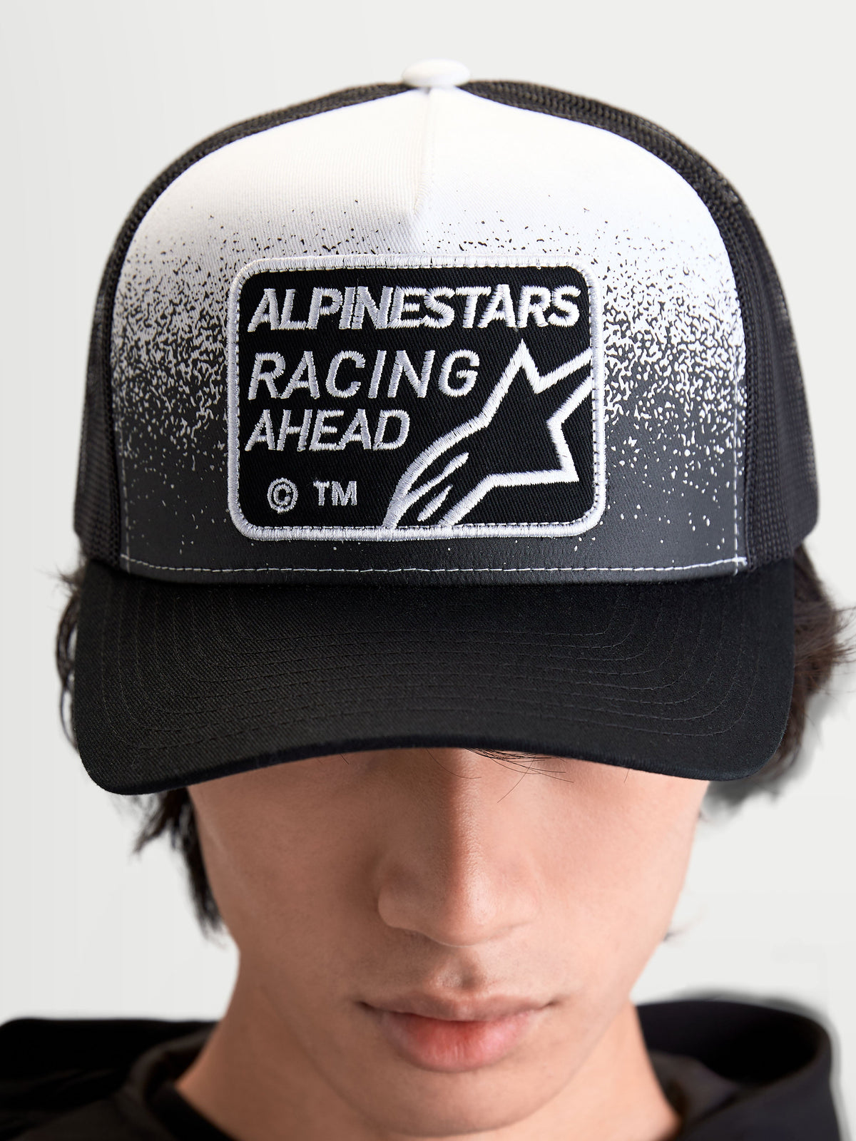 Alpinestars Gravel Trucker Hat, Sportswear Trucker Cap for Men and Women, Black and White, featuring a woven Alpinestars Racing Ahead patch with gravel-texture detailing, structured crown, and curved bill, on-model front view.