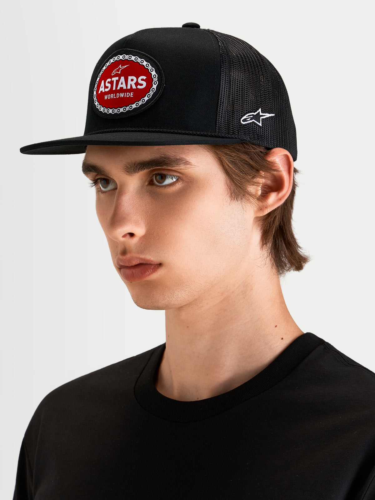 Alpinestars Hotlink Trucker Hat, motorsport-inspired flat bill snapback cap worn by a model, black and red, featuring a structured crown and front Astars logo patch with bike chain border