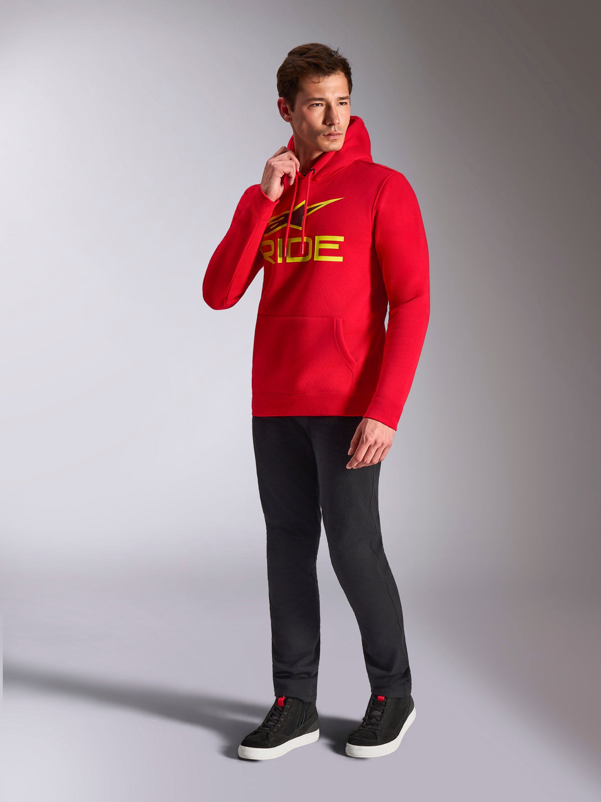 Alpinestars Ride V3 Hoodie, red pullover sweatshirt, yellow and black 'RIDE' logo on chest, worn by model with black pants and black sneakers, casual motorsport-inspired streetwear