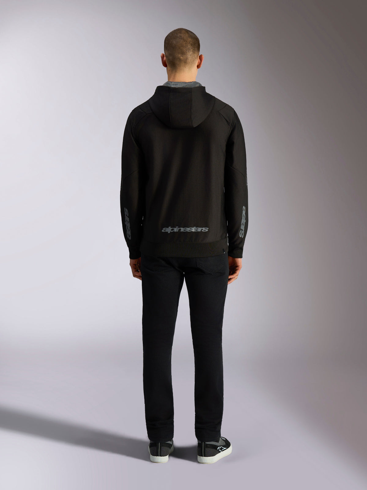 Back view of Alpinestars Sessions V3 Hoodie in Black Grey, featuring a grey Alpinestars wordmark on the lower back and astars logos on the forearms, worn by a model with black pants, casual motorsport-inspired streetwear