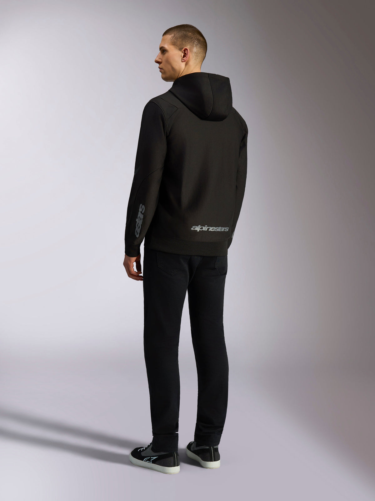 Alpinestars Sessions V3 Hoodie, black casual streetwear, back view on a model, featuring grey alpinestars wordmark on the lower back and Astars logo on the sleeve, hooded zip-front design with modern motorsport-inspired styling