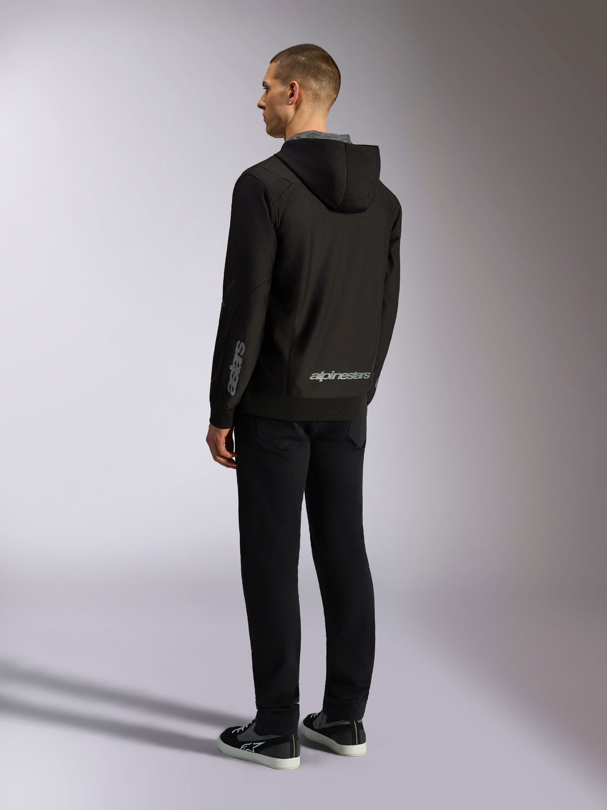 Alpinestars Sessions V3 Hoodie, casual motorsport streetwear, black with grey wordmark on the lower back and forearm, zip-front poly fleece with mesh-lined hood, back view on model, modern athletic design