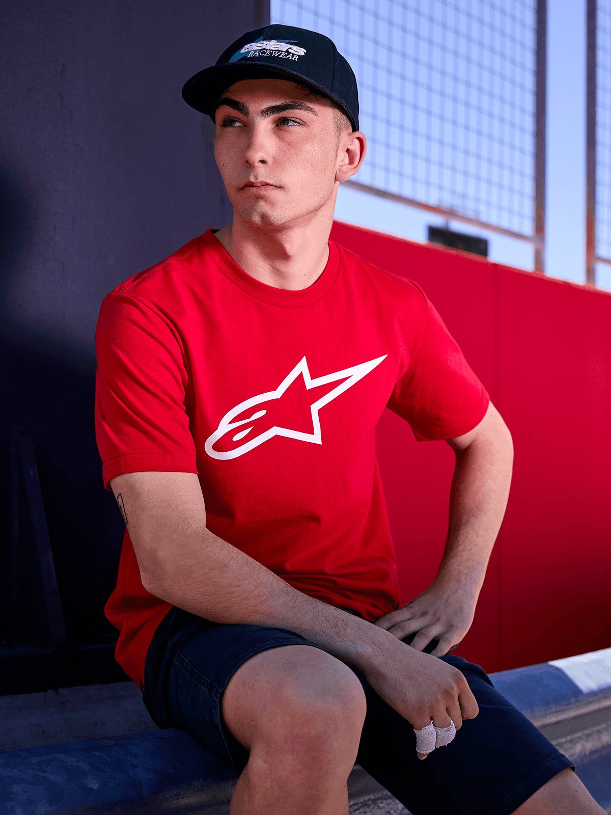 Alpinestars Profile Hat in Black, lifestyle shot of a man wearing the curved bill hat with a red t-shirt featuring a white Alpinestars logo, casual motorsport-inspired streetwear