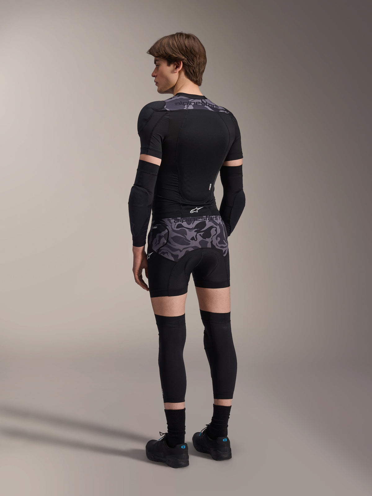 Model wearing Alpinestars A-Motion Plasma Protection Boxers in Black Camo and matching A-Motion Plasma Protection Jacket, rear view showing grey camouflage pattern, elbow guards, knee protectors, and cycling shoes, complete mountain bike protection gear setup.