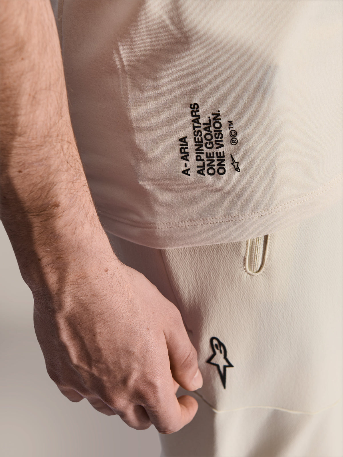 Alpinestars A-Aria Polartec Elite Short Sleeve Jersey, Ecru color, close-up of side branding featuring 'A-ARIA', 'ALPINESTARS', 'ONE GOAL. ONE VISION.', and Astars logo, worn with matching off-white pants, premium MTB performance apparel
