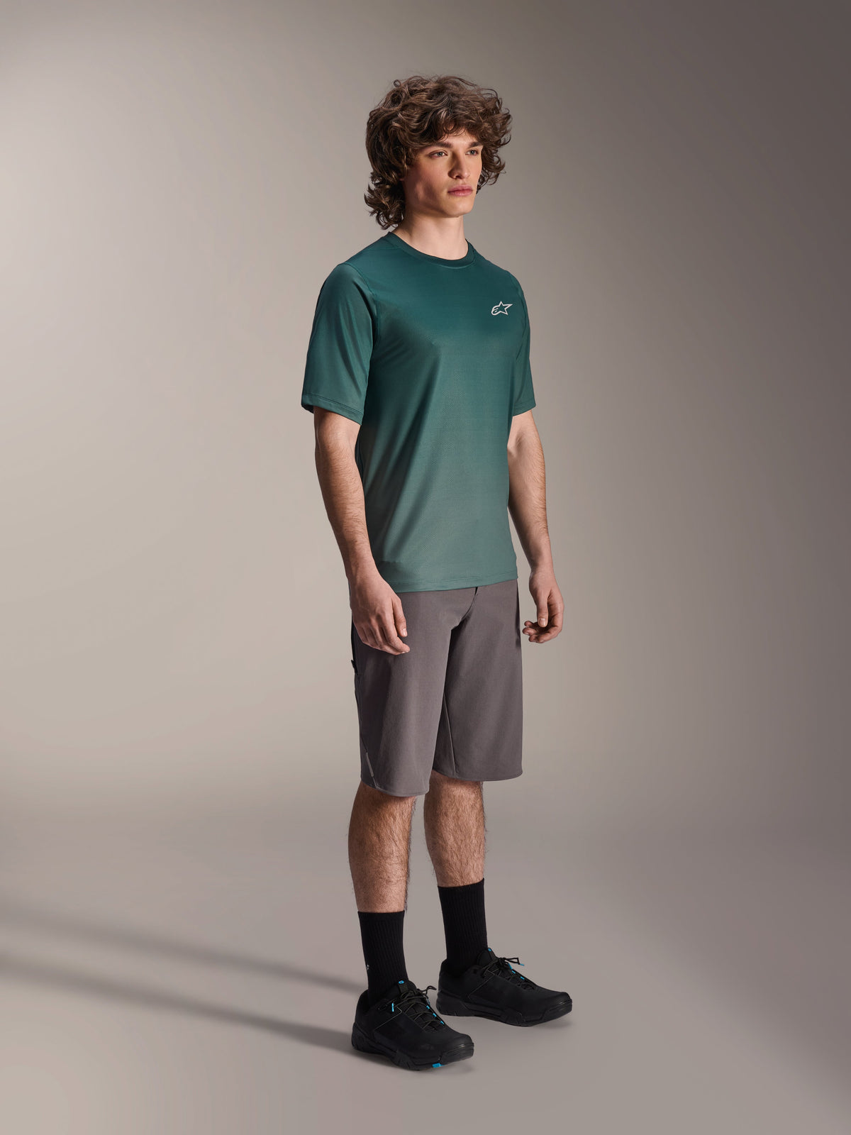 Alpinestars A-Dura Rise Jersey - Short Sleeve in Silver Pine, green mountain bike jersey worn by a model with grey shorts and black sneakers, casual MTB trail riding gear featuring a small white chest logo