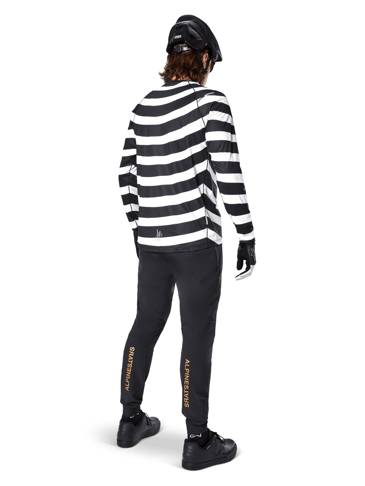 MTB rider wearing the Alpinestars A-Aria Oscar Long Sleeve Jersey, black and white horizontal spiral stripes, paired with black technical pants featuring gold logos on the calves, black helmet and shoes, complete off-road cycling outfit from behind