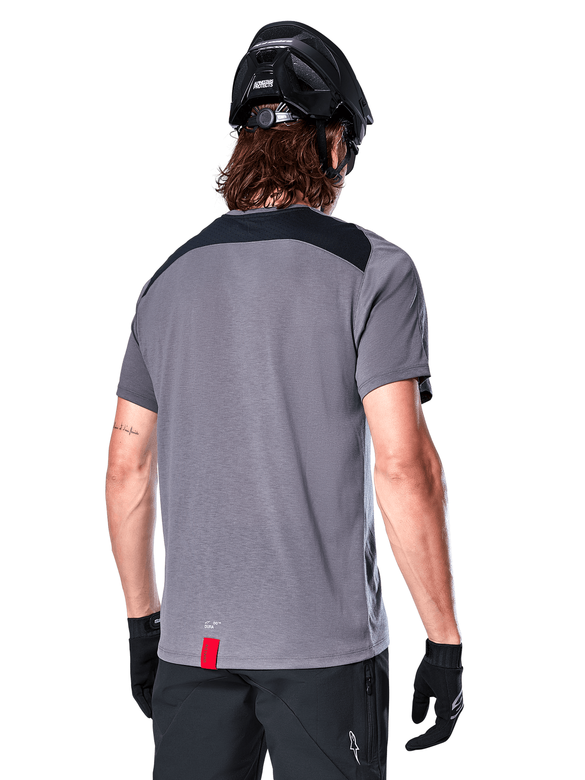 Alpinestars A-Dura Dri Astar Short Sleeve Jersey in Dark Gray, back view showing black mesh panels on the upper back and a longer drop tail, worn with a black MTB helmet and black shorts, technical mountain bike apparel.