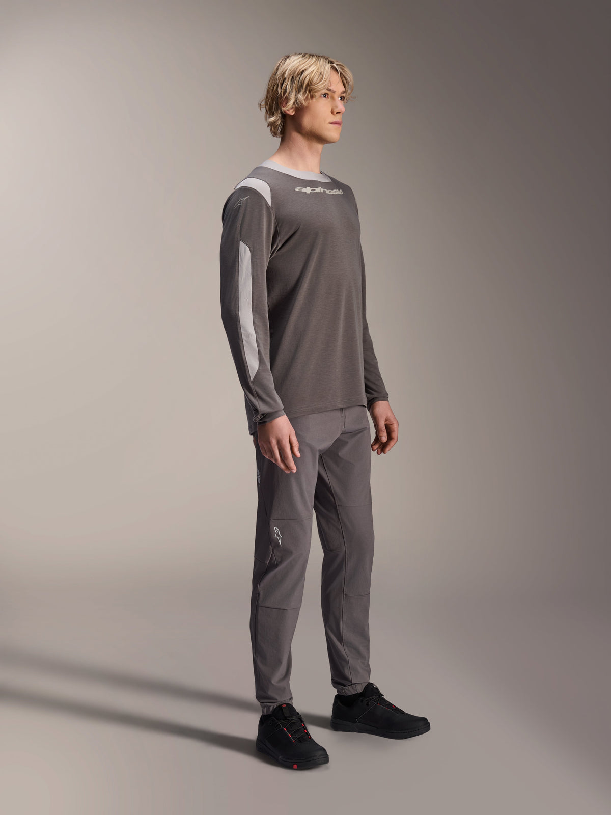 Male model wearing Alpinestars A-Dura Dri Wool Long Sleeve Jersey in Dark Gray, paired with matching gray pants and black shoes, featuring light gray mesh panels on the shoulders and sleeves, technical MTB gear for off-road cycling