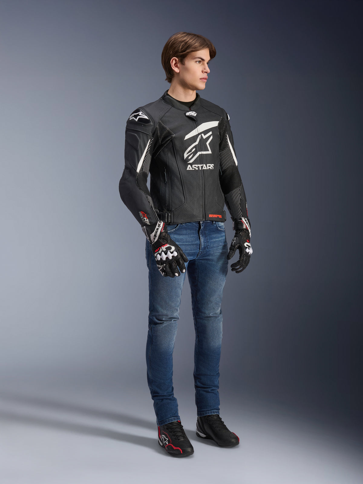 Alpinestars GP Plus R V4 Rideknit Leather Jacket, Black and White, motorcycle leather jacket worn by a model with blue jeans and black gloves, featuring a prominent white Astars logo on the chest, perforated panels, and white accents on the shoulders.