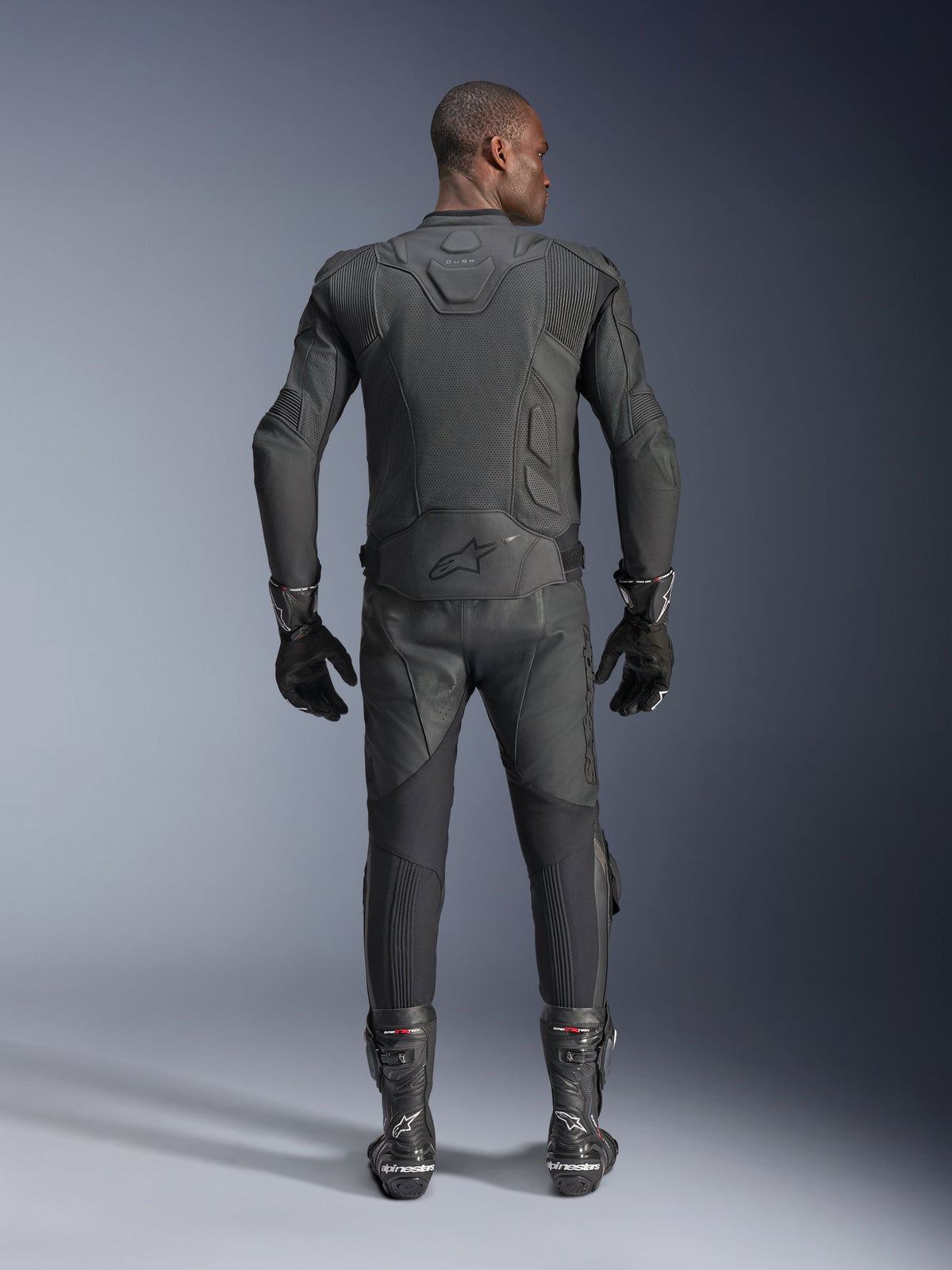 Male model shown from the back wearing the Alpinestars Dusk Airflow Leather Jacket in Black, paired with matching black leather racing pants, gloves, and boots, featuring extensive perforations for ventilation and the Alpinestars logo on the lower back.