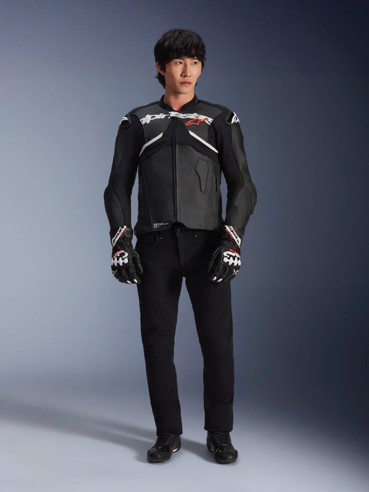 Model wearing the Alpinestars Atem V5 Leather Jacket in Black and White, featuring white Alpinestars branding and a red star logo on the chest, paired with black pants and black and white racing gloves, sleek performance motorcycle gear