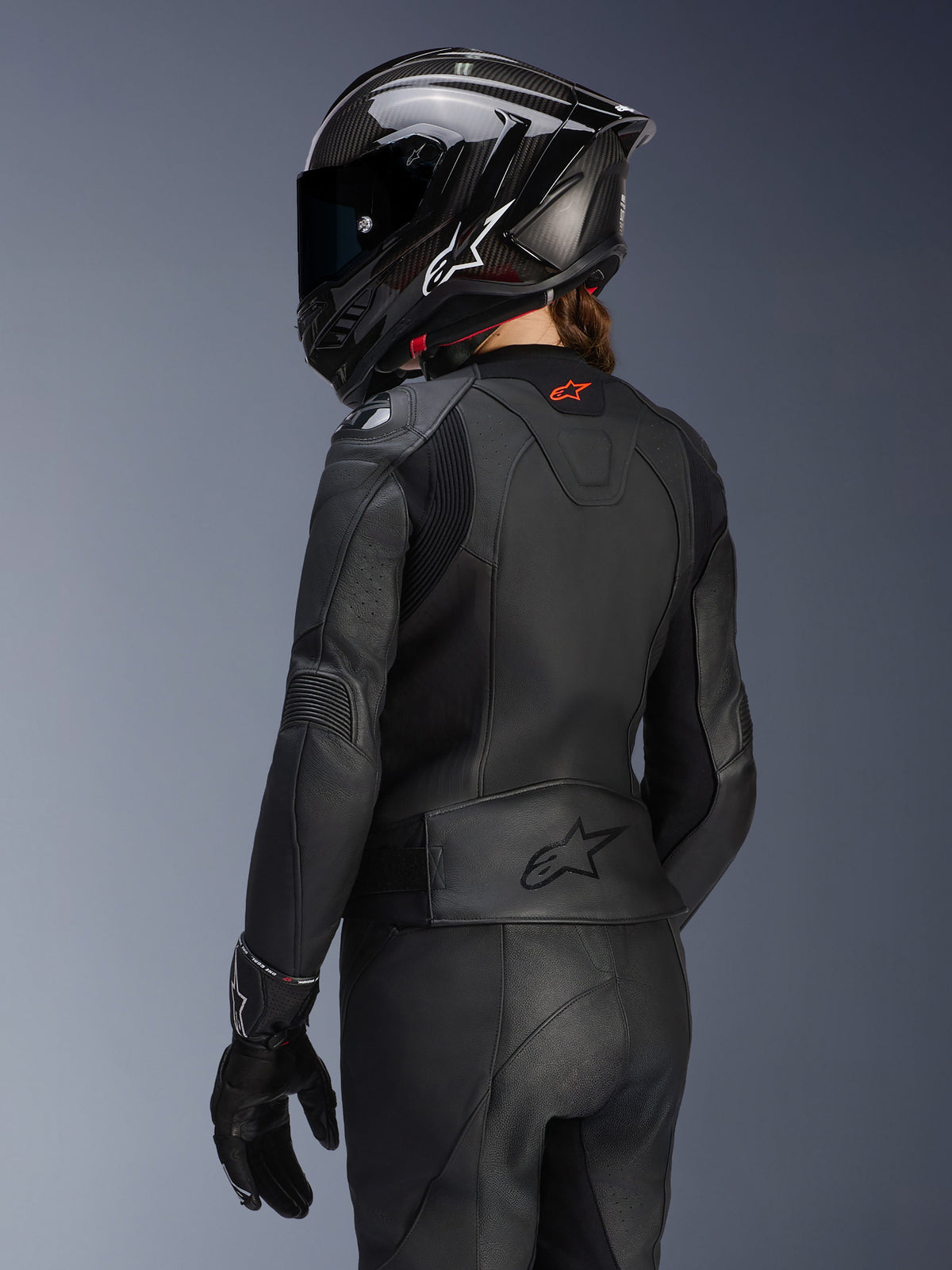 Female rider from the back wearing the Alpinestars Women's Stella GP Plus V4 Leather Jacket in Black, matching leather pants and gloves, and a black carbon-style full-face helmet, highlighting the jacket's aerodynamic fit and embossed logo on the lower back.