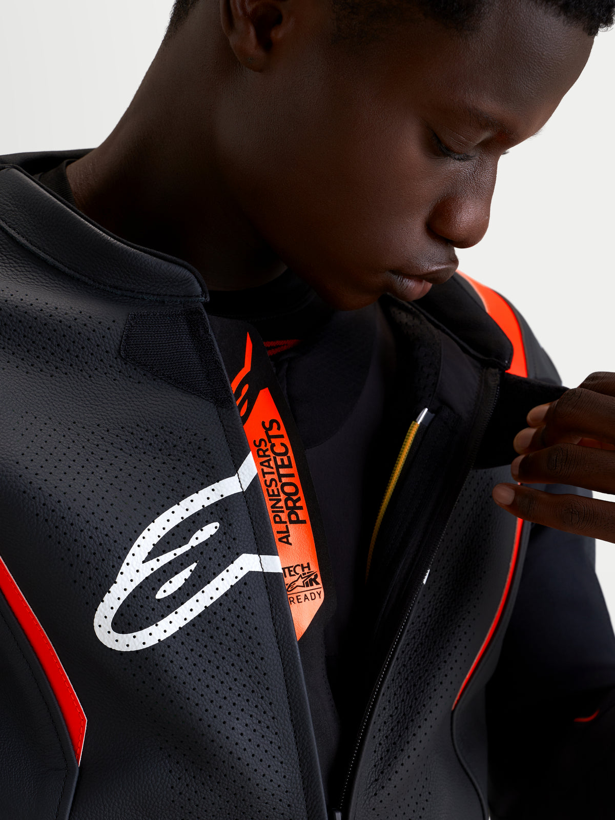 Alpinestars Missile V3 Leather Suit - 2 Piece, close-up of a model wearing the black and red fluo racing leather suit, showing the perforated front closure tab with white Astar logo and Alpinstars Protects branding