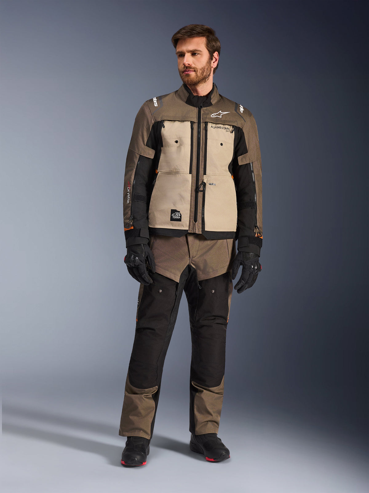 Alpinestars Cusco Drystar® Jacket in Walnut Aluminum Black, worn by a male rider with matching adventure touring pants, black gloves, and boots, featuring tan chest panels, black sleeves with subtle orange accents, and functional ventilation zippers for motorcycle road riding.