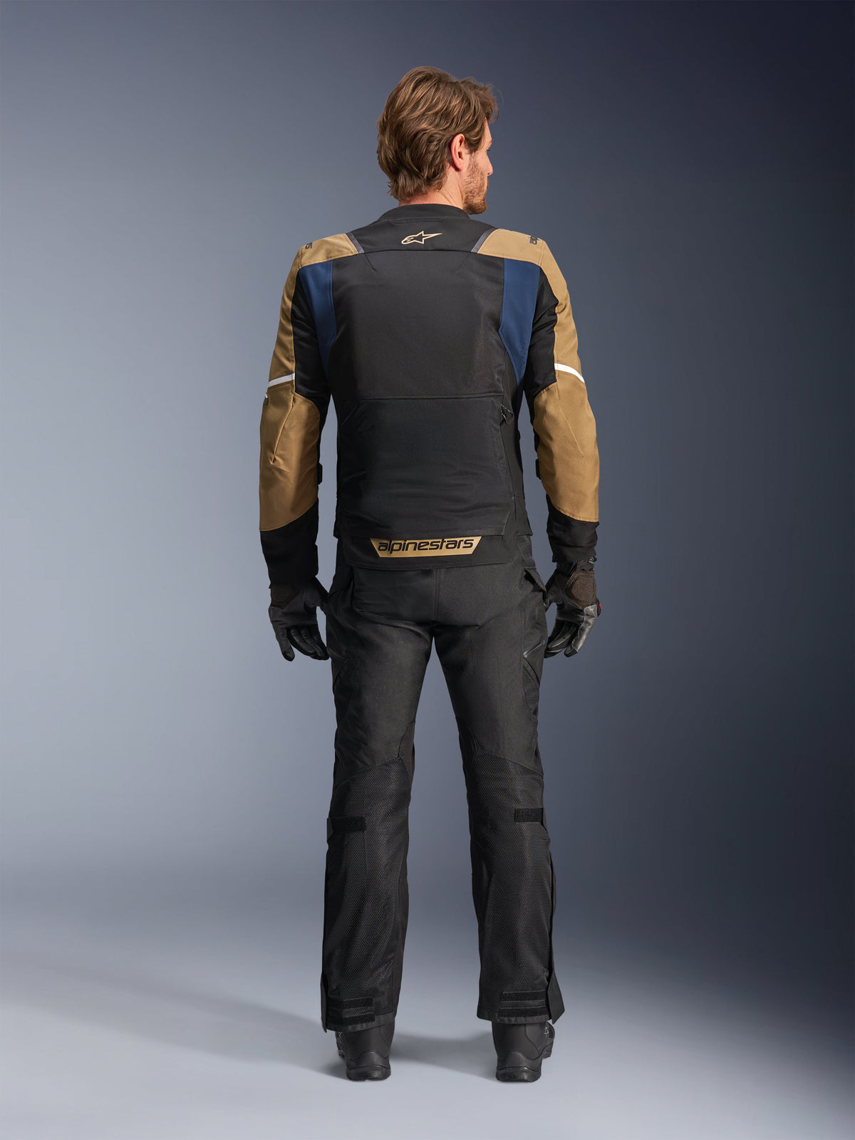 Alpinestars ST-2 Air Jacket in Deep Blue, Dark Khaki, and Black, rear view on model showing large mesh back panel and khaki sleeves with white stripes, paired with black technical motorcycle pants and gloves for adventure touring.