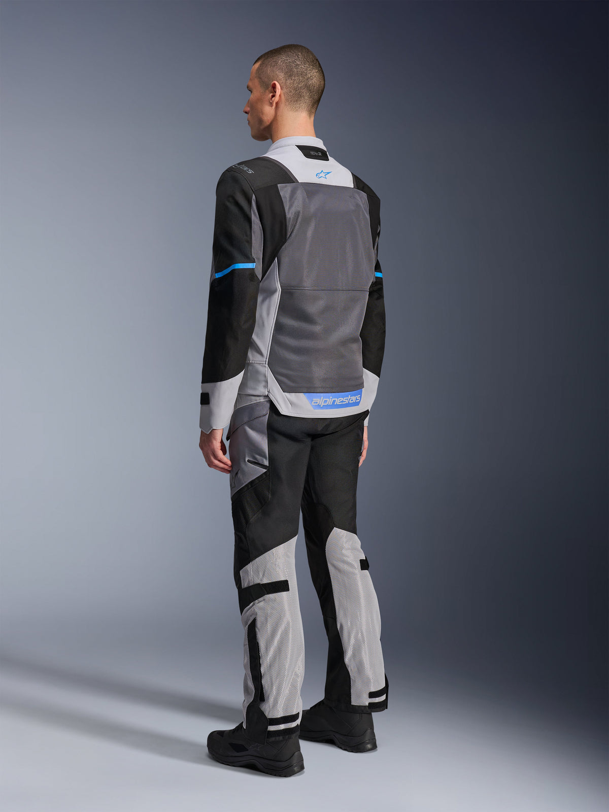 Male model from behind wearing the Alpinestars ST-2 Air Jacket in Ice Gray, Dark Gray, and Black, featuring a large grey mesh back panel, blue logo accents, and matching motorcycle pants for summer touring.