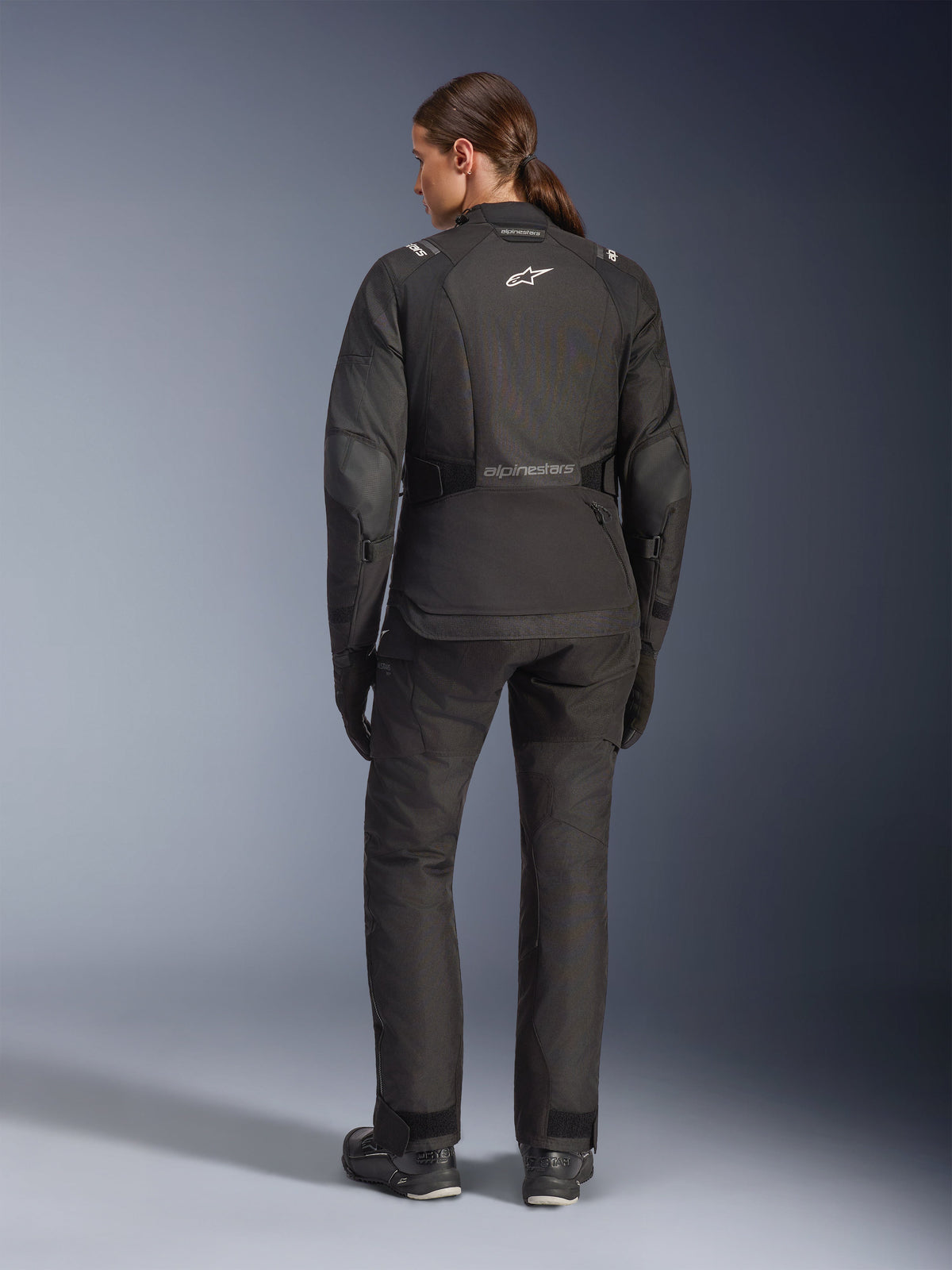 Female rider from behind wearing the Alpinestars Stella Andes V4 Drystar® Jacket, Black, paired with matching motorcycle pants, white Astar logo on the back, elongated rear hem, complete adventure-touring riding outfit including black gloves and boots