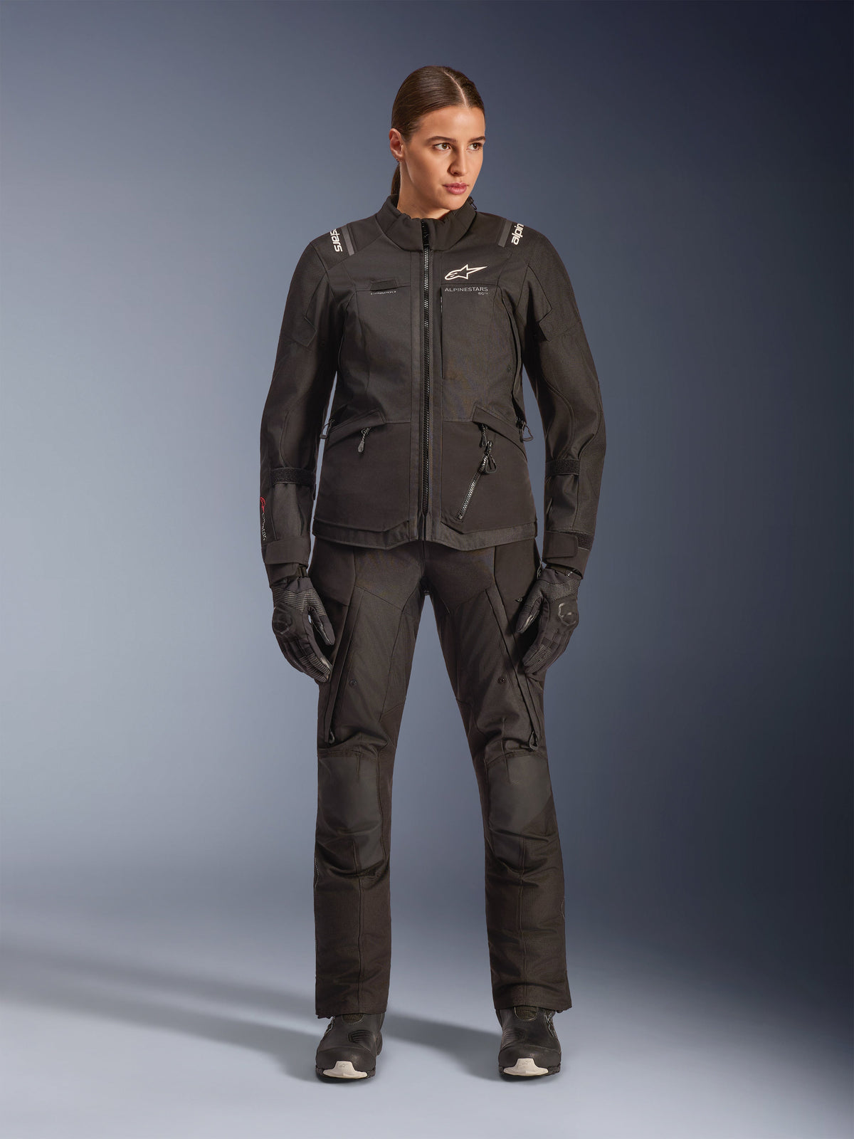 Female rider wearing Alpinestars Stella Andes V4 Drystar® Jacket in Black, paired with matching black adventure-touring pants and gloves, featuring technical textile construction with multiple utility pockets and white Alpinestars branding, complete all-weather motorcycle gear