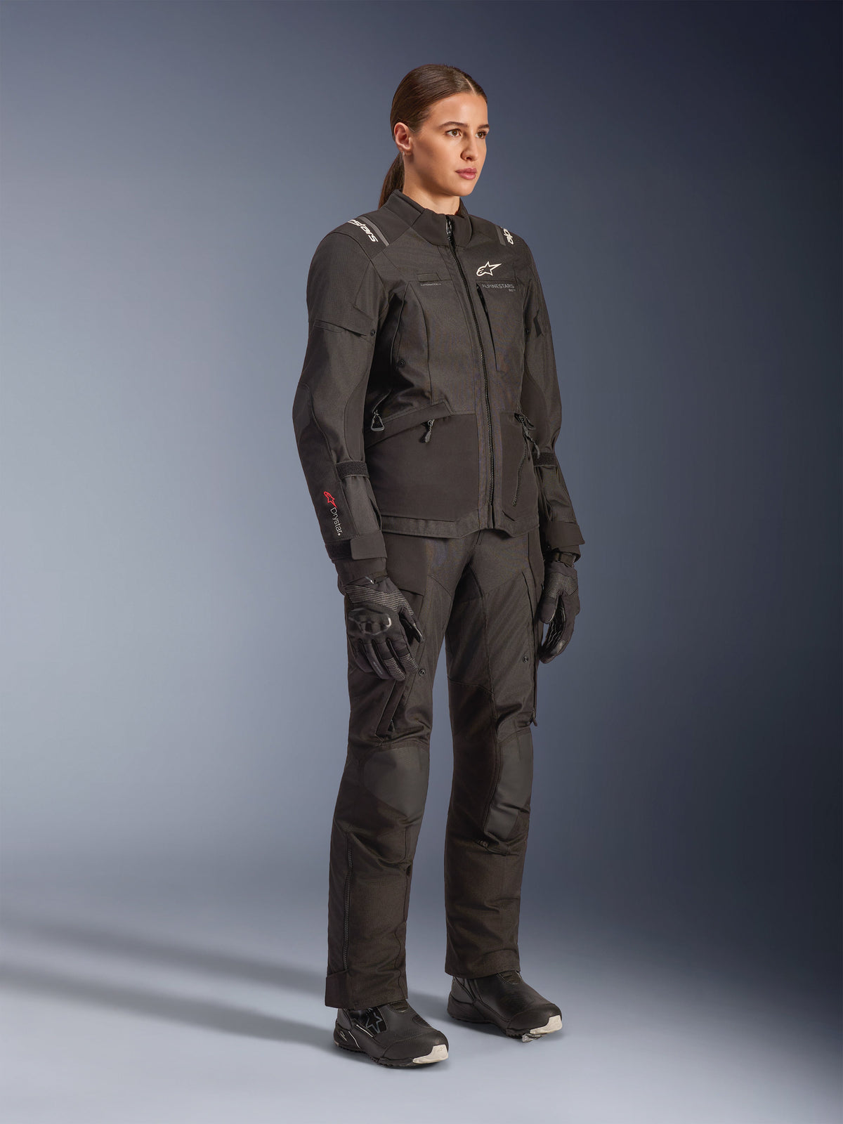 Female rider wearing the Alpinestars Woman Stella Andes V4 Drystar® Jacket in black, paired with matching adventure touring pants, gloves, and boots, featuring white Alpinestars logos and red Drystar branding, designed for all-weather motorcycle performance.