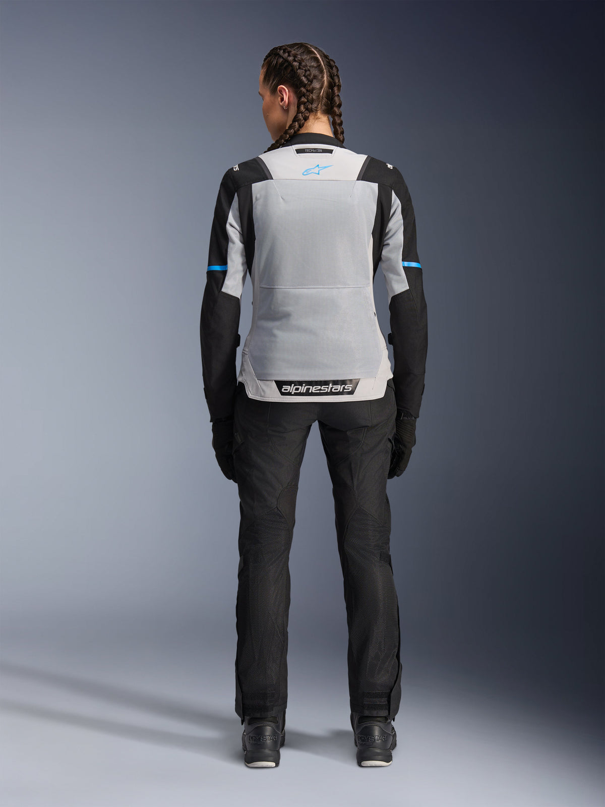 Alpinestars Stella ST-2 Air Jacket, Ice Gray Black, rear view on female model, featuring a large light gray mesh back panel, black sleeves with blue stripe accents, Alpinestars branding on the lower back, paired with black technical riding pants and boots, summer motorcycle touring gear