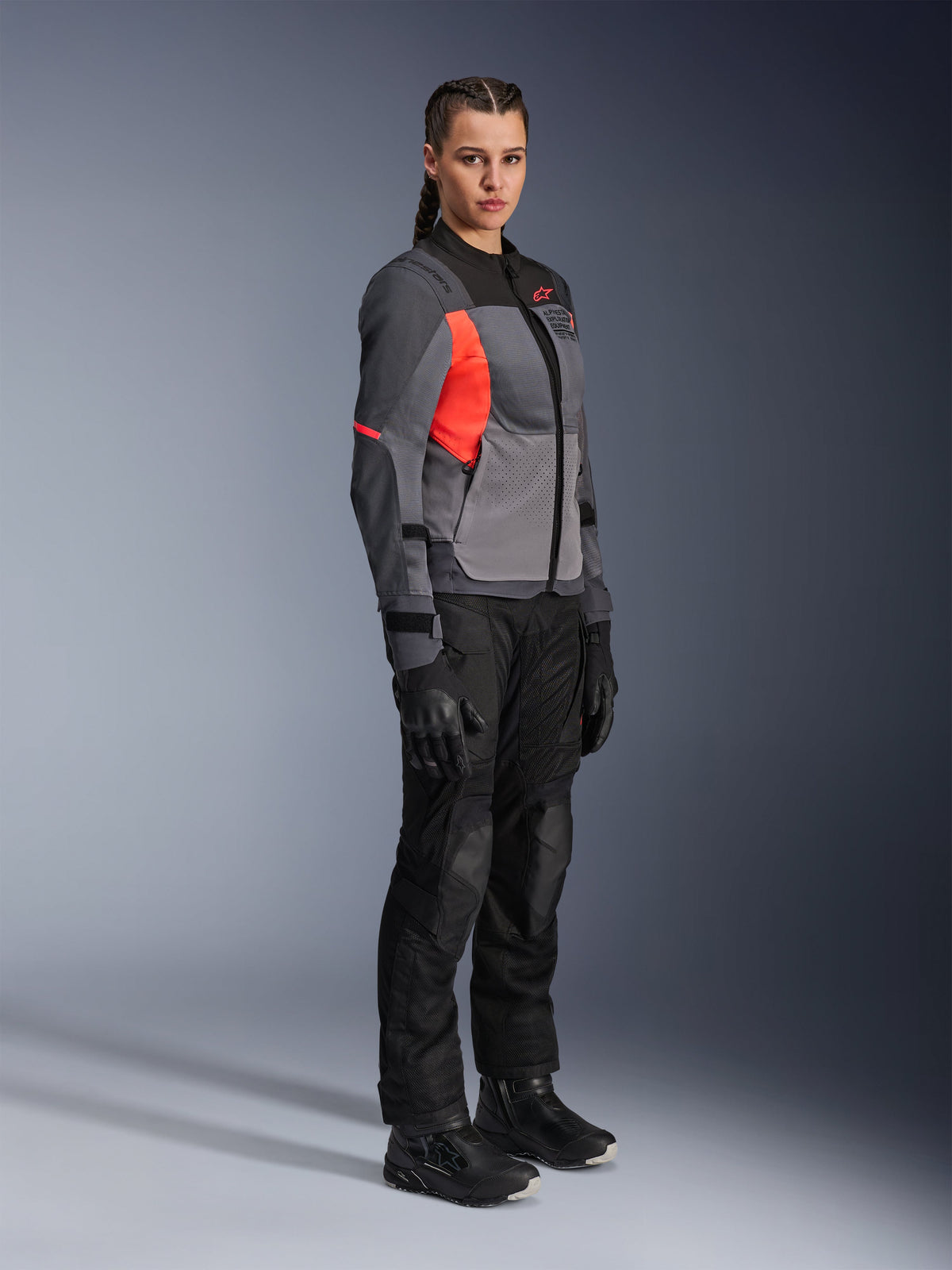 Woman rider wearing the Alpinestars Stella ST-2 Air Jacket in Deep Gray, Dark Gray, and Fluo Coral, paired with black motorcycle pants, gloves, and boots, featuring a modern touring design with mesh panels and reflective details.