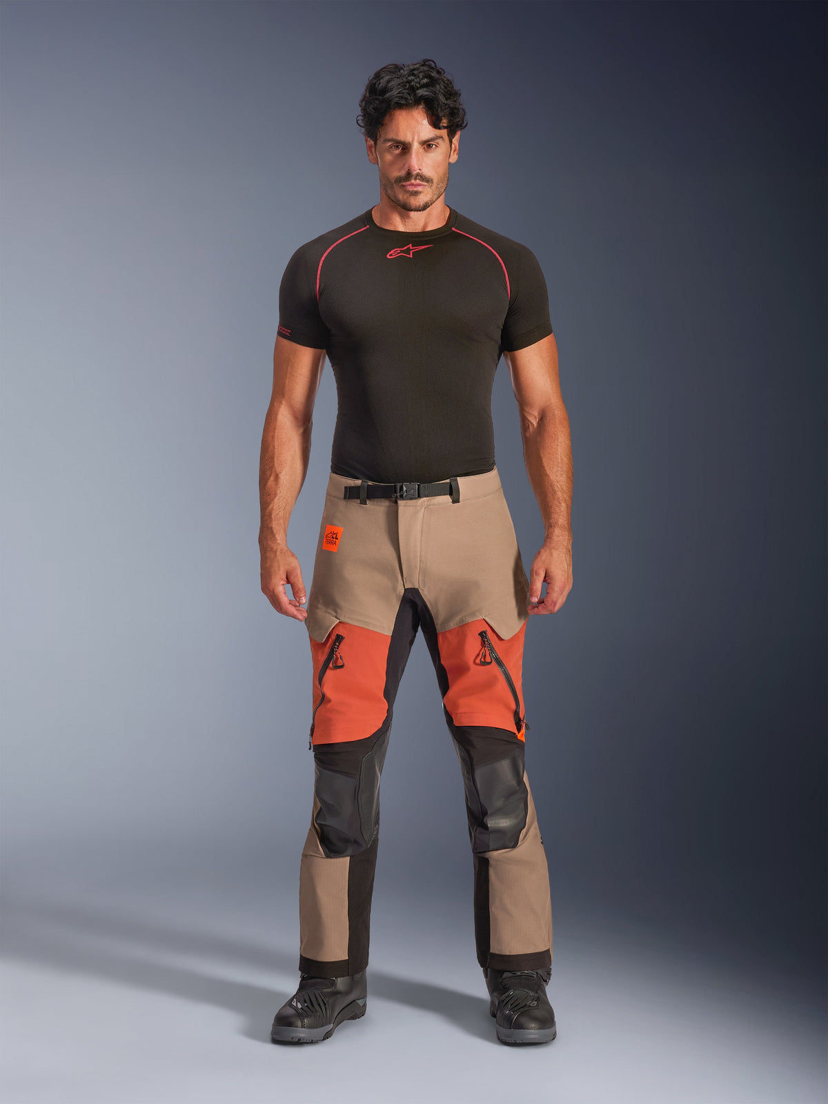Male model wearing Alpinestars Halo Pro Drystar® XF Laminated Pants, Adventure Motorcycle Pants, Walnut Rust Black, tan and orange color-blocked design with black technical panels, featuring zippered thigh vents and magnetic waist buckle, paired with a black base layer and black motorcycle boots