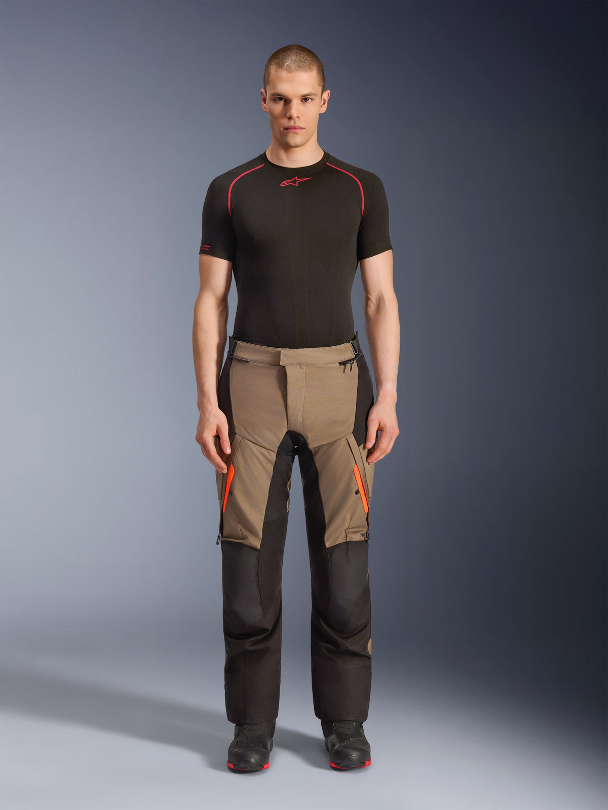 Male model wearing Alpinestars Andes V4 Drystar® Pants in Walnut Black, adventure motorcycle pants paired with a black base layer and boots, featuring orange zipper accents on the thighs and reinforced black panels for all-terrain durability.