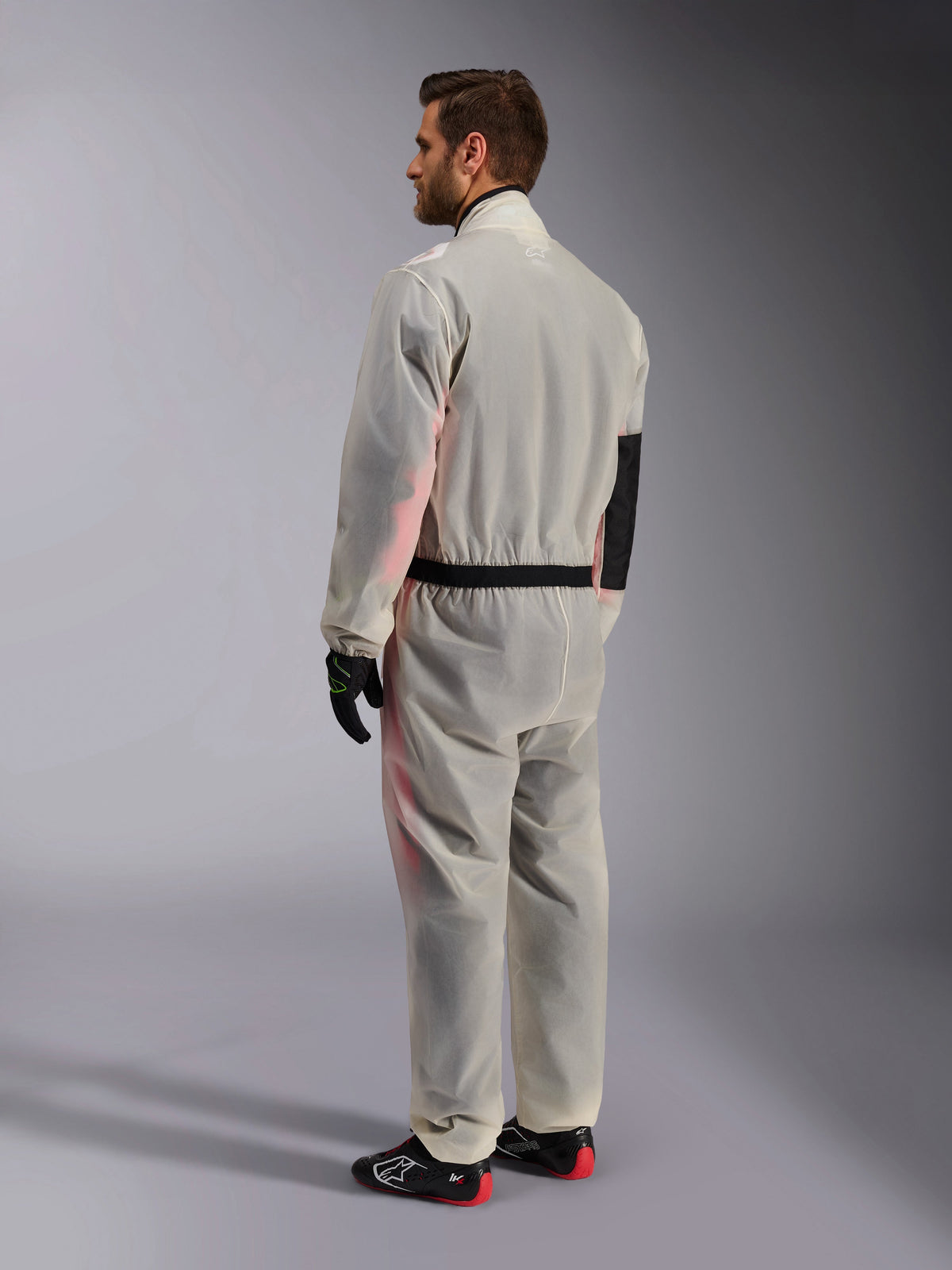 Alpinestars Kart Rain Suit, transparent waterproof karting oversuit, Clear, shown from a rear three-quarter view, featuring a black reinforced elbow panel and elasticated waistband, worn over a racing suit with black and red shoes, designed for wet weather karting